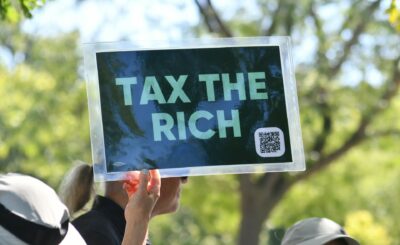Person që mban pankartë me mesazhin 'Tax the Rich' gjatë një proteste për politikat tatimore