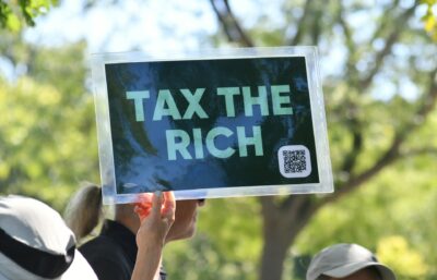 Person që mban pankartë me mesazhin 'Tax the Rich' gjatë një proteste për politikat tatimore