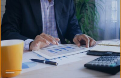 A person in a suit is looking at financial charts on a tablet next to a calculator and a notepad.