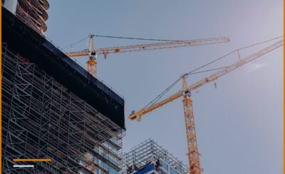 Due gru a torre arancioni sopra un edificio in costruzione, viste dal basso, con le impalcature della facciata in primo piano.