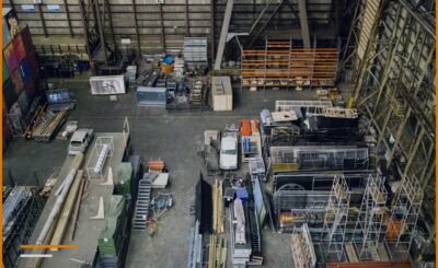 Una vista dall'alto di un capannone di produzione, con scaffali arancioni, materiali in legno e metallo e strutture in fase di lavorazione sparse sul pavimento.