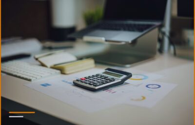 A work desk with a calculator, a notepad, and printed reports next to a monitor.
