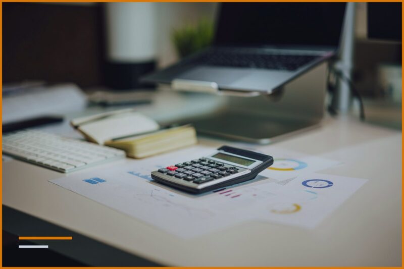 A work desk with a calculator, a notepad, and printed reports next to a monitor.